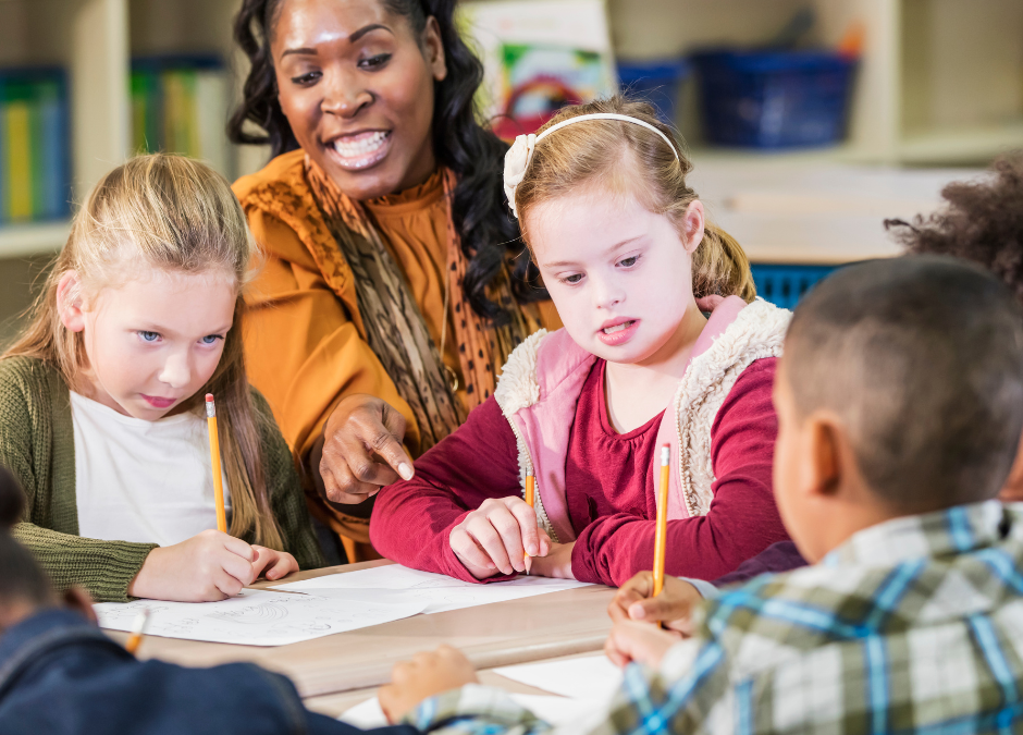 a teacher pointing at the table where four students are sitting and writing with pencils on paper