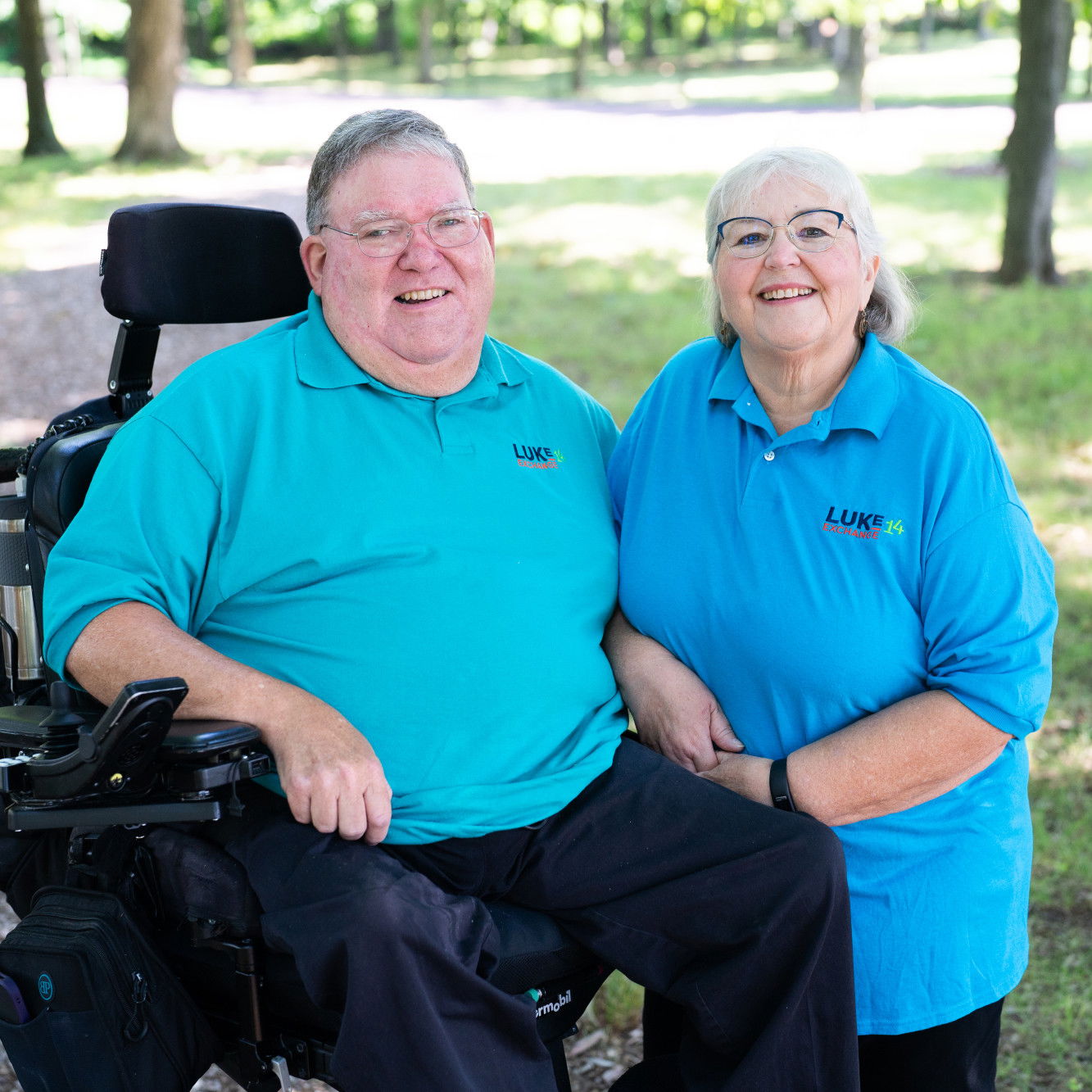 Jerry in a wheelchair with Joan beside him.  They are outside.  Shadows are spotty in the grass behind them.  They are both wearing blue polo shirts.