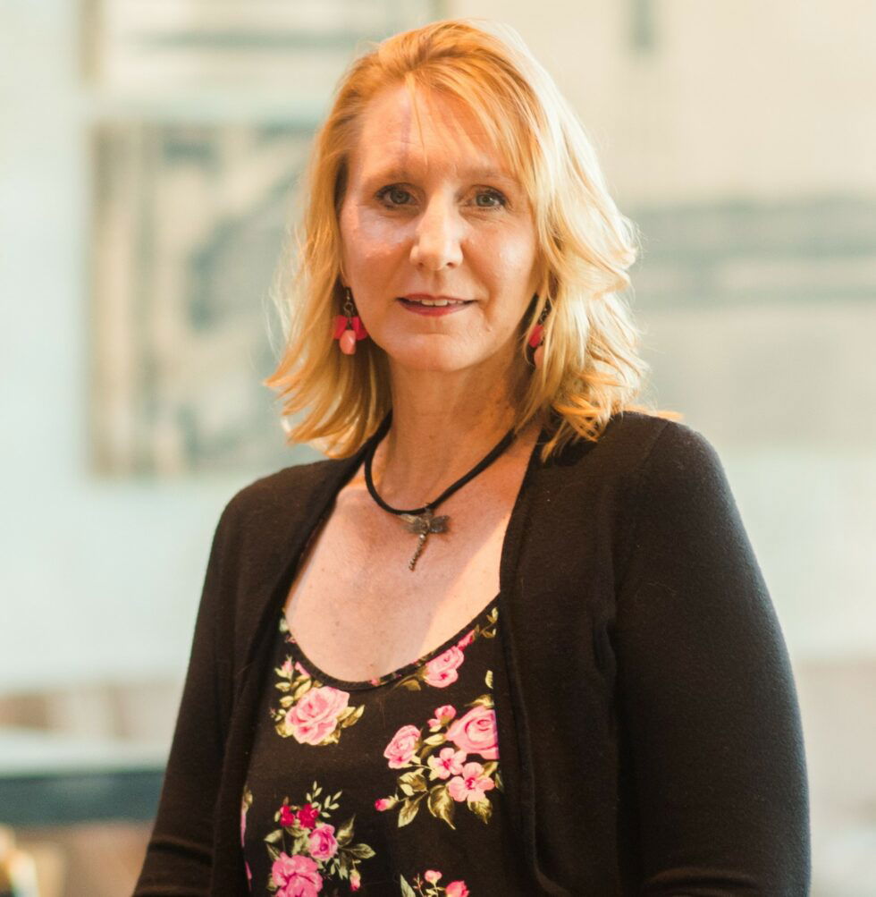 Woman with blonde hair wearing a shirt with pink roses and a black cardigan.