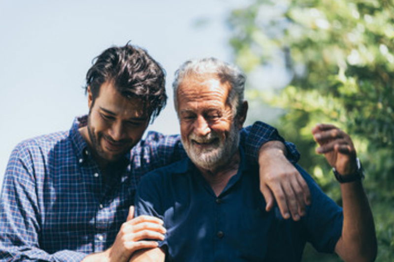 Man and older dad outside and smiling.  Son's arm is around his father's shoulder.